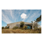 A rectangular art print showing a modern concrete building nestled in dry grass under a bright blue sky with wispy white clouds from the hardcover book "contemporary living yearkbook 2026" on a white background