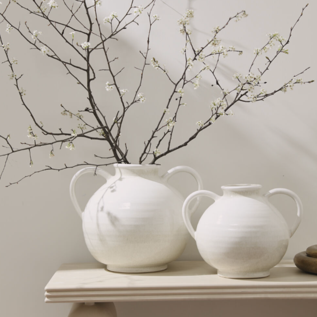 Two white ceramic vases with handles sit on a light wood table, one filled with flowering branches against a neutral beige background.