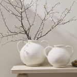 Two white ceramic vases with handles sit on a light wood table, one filled with flowering branches against a neutral beige background.