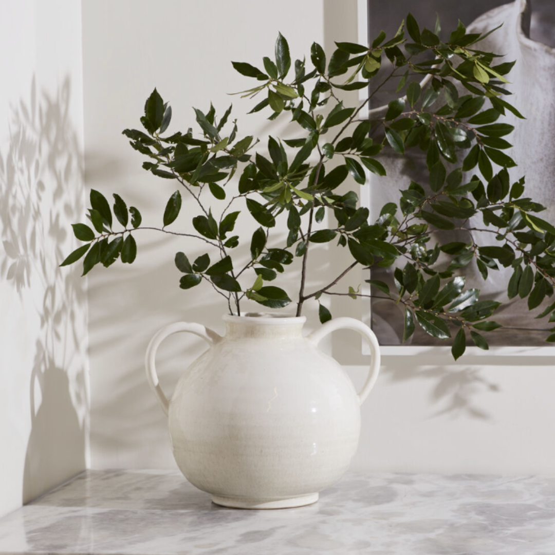 A white ceramic double-handled vase holding green leafy branches, set on a light marble surface against a white wall with soft shadows.