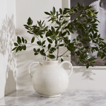 A white ceramic double-handled vase holding green leafy branches, set on a light marble surface against a white wall with soft shadows.