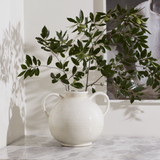 A white ceramic double-handled vase holding green leafy branches, set on a light marble surface against a white wall with soft shadows.
