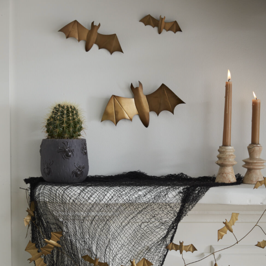 a spooky decorated mantel with two candlesticks and taper candles, black netting, gold bat garlands, potted green plant, and three gold bats on the wall. 