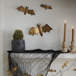 a spooky decorated mantel with two candlesticks and taper candles, black netting, gold bat garlands, potted green plant, and three gold bats on the wall. 