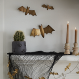 a spooky decorated mantel with two candlesticks and taper candles, black netting, gold bat garlands, potted green plant, and three gold bats on the wall. 