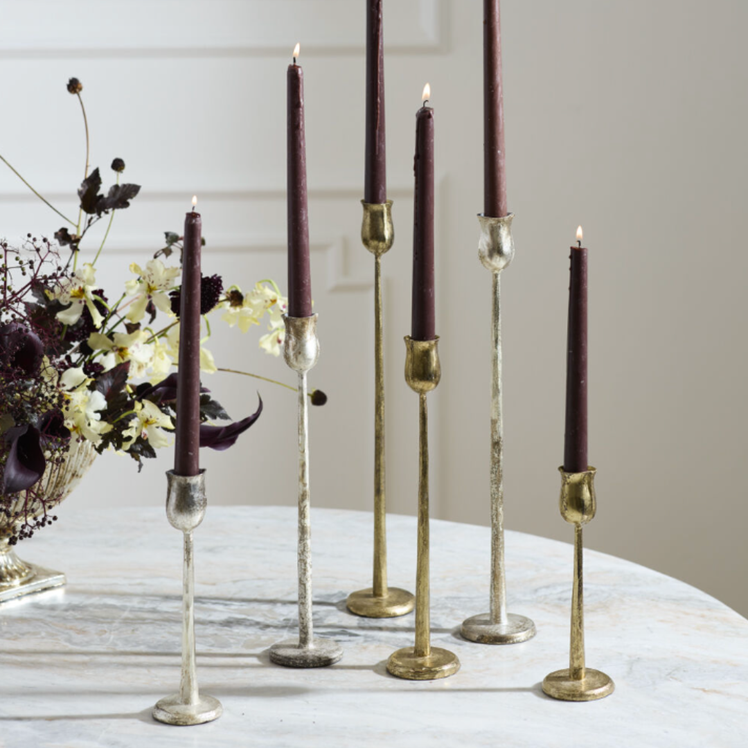 Five taper candles in dark burgundy or plum tones displayed in mixed metallic candlesticks on a light marble surface, with dried flowers in the background.