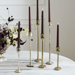 Five taper candles in dark burgundy or plum tones displayed in mixed metallic candlesticks on a light marble surface, with dried flowers in the background.