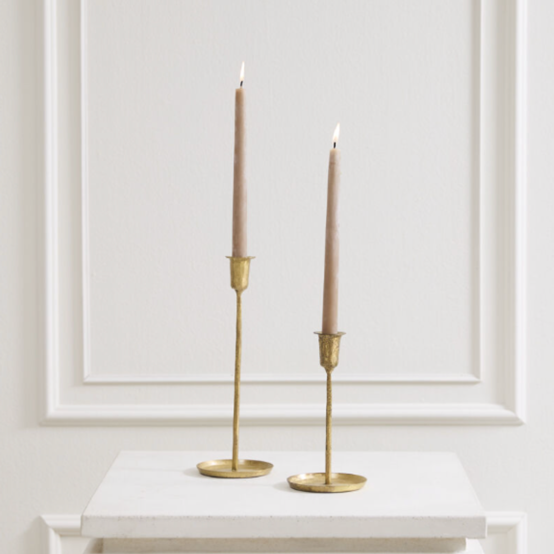 Two slim beige taper candles in gold metal candlesticks, displayed on a white marble-topped surface against a white paneled wall.