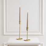 Two slim beige taper candles in gold metal candlesticks, displayed on a white marble-topped surface against a white paneled wall.