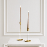 Two slim beige taper candles in gold metal candlesticks, displayed on a white marble-topped surface against a white paneled wall.