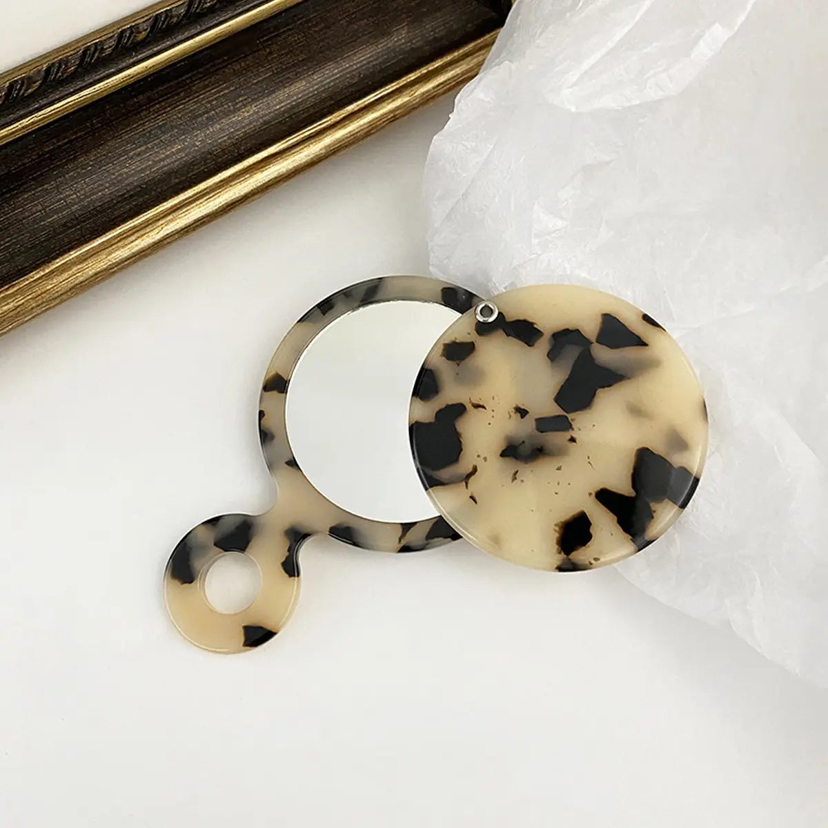Tortoiseshell compact mirror on a white surface with a gold frame in the background.