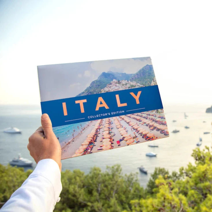 Person holding a travel guidebook titled 'Italy' with a scenic beach in the background.