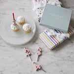Cupcakes with pink number candles on a marble surface, next to gift boxes and colorful striped fabric.