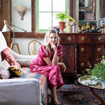 Woman in a pink dress sitting on a sofa in a decorated room with wooden furniture and plants.