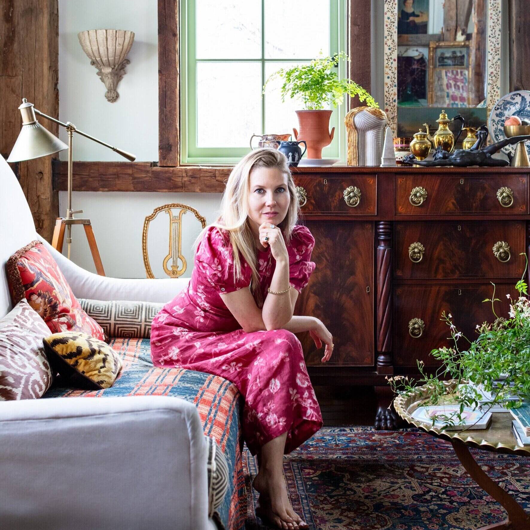 Woman in a pink dress sitting on a sofa in a decorated room with wooden furniture and plants.