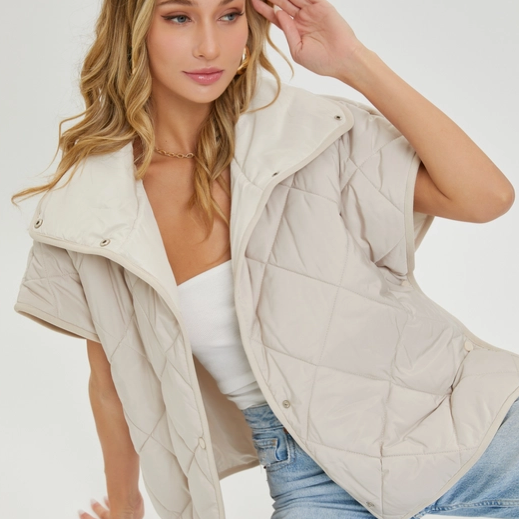 Model wearing a taupe quilted short-sleeve jacket with a high collar and relaxed fit, paired with light blue jeans and a white tank top."