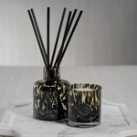 Decorative diffuser with black reeds and a candle in a glass holder on a white surface.