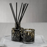 Decorative diffuser with black reeds and a candle in a glass holder on a white surface.