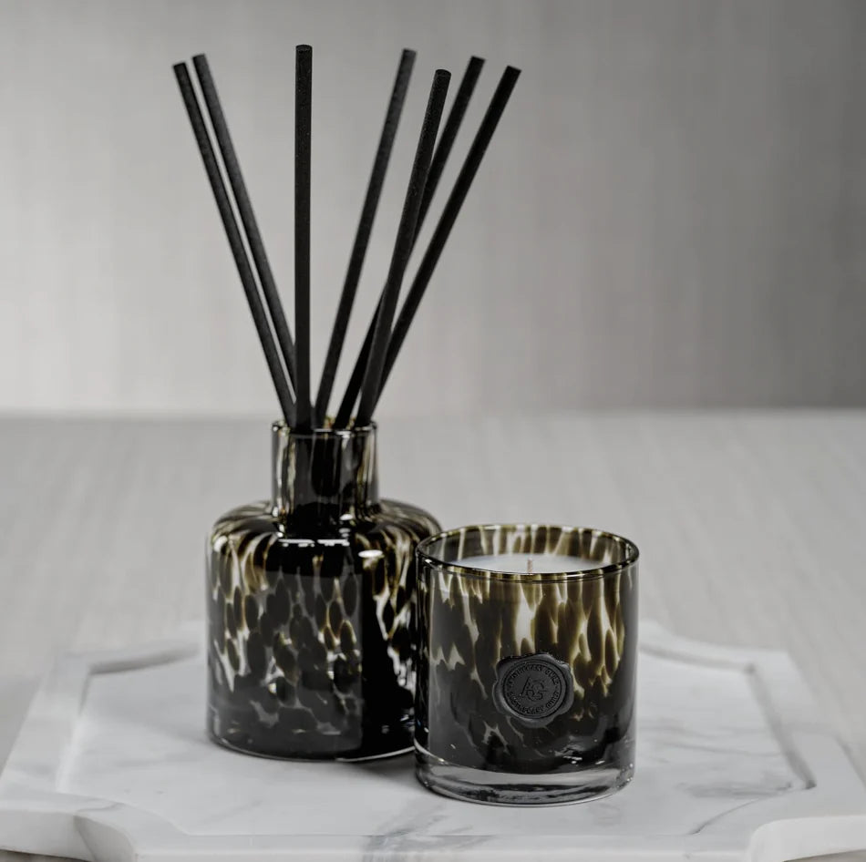 Decorative diffuser with black reeds and a candle in a glass holder on a white surface.