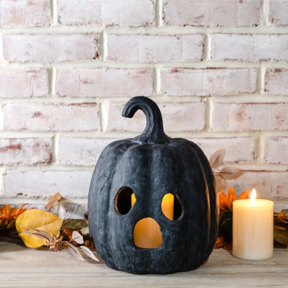 Black pumpkin-shaped candle holder with a lit candle against a white brick wall.