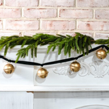 Decorative garland with gold bells on a white fireplace against a brick wall.