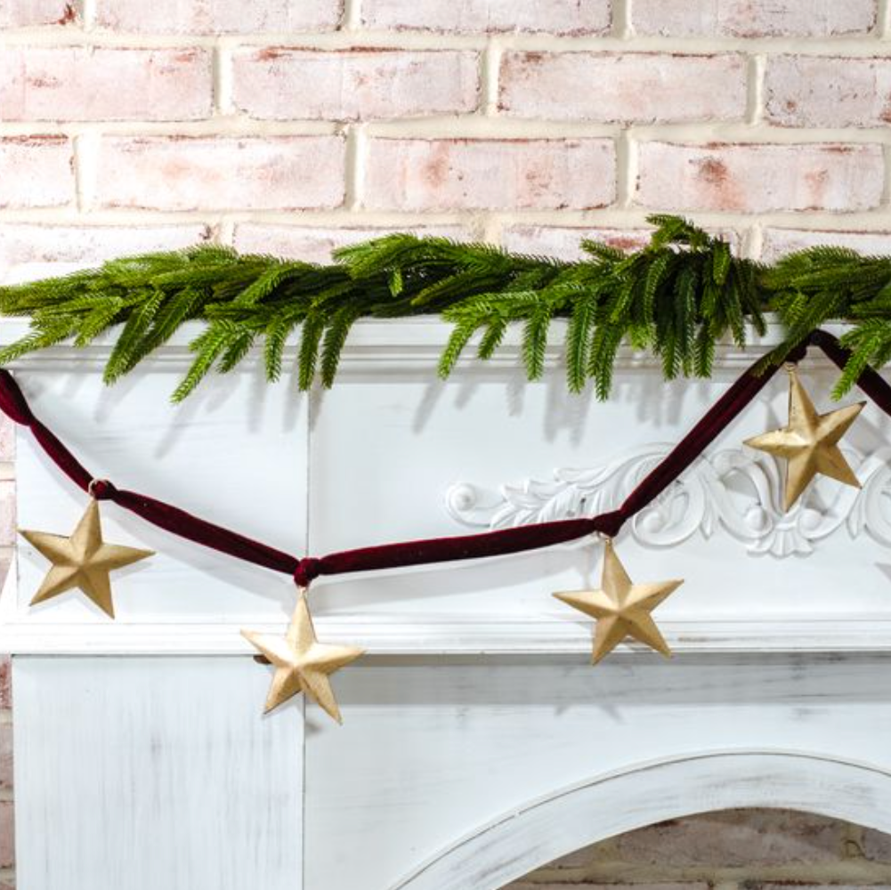 Decorative Christmas mantel with greenery, gold stars, and red ribbon against a white brick wall.