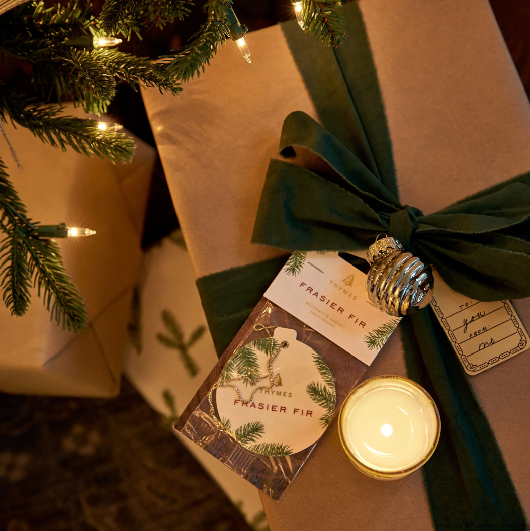 Gift wrapped in brown paper with green ribbon and tag, next to a lit candle under a Christmas tree.