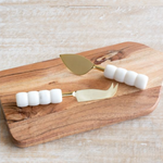 Wooden cutting board with gold cheese knives and white cheese on a light wooden surface