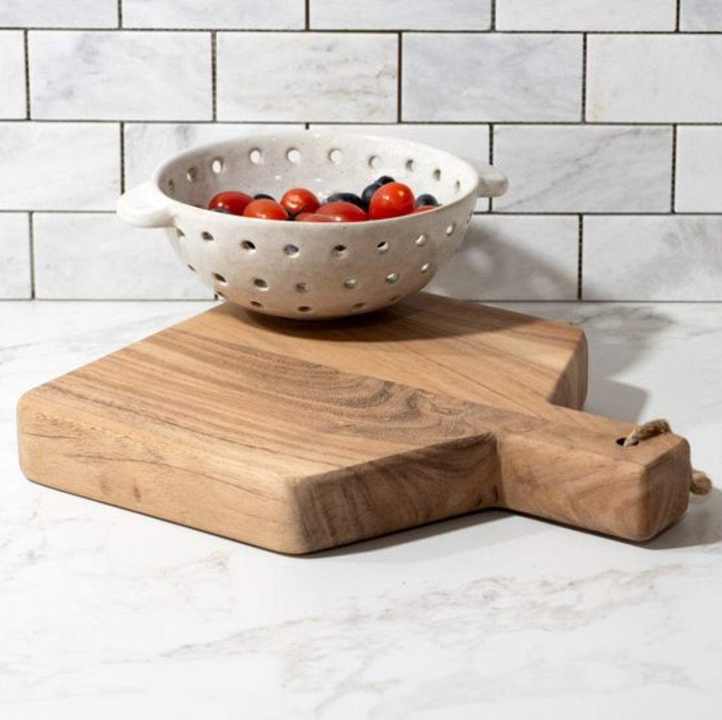 Wooden cutting board with a colander containing tomatoes on a marble countertop.