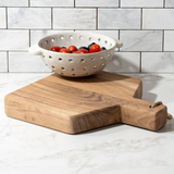Wooden cutting board with a colander containing tomatoes on a marble countertop.