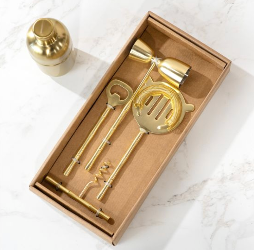 Gold cocktail shaker and tools in a cardboard box on a marble surface