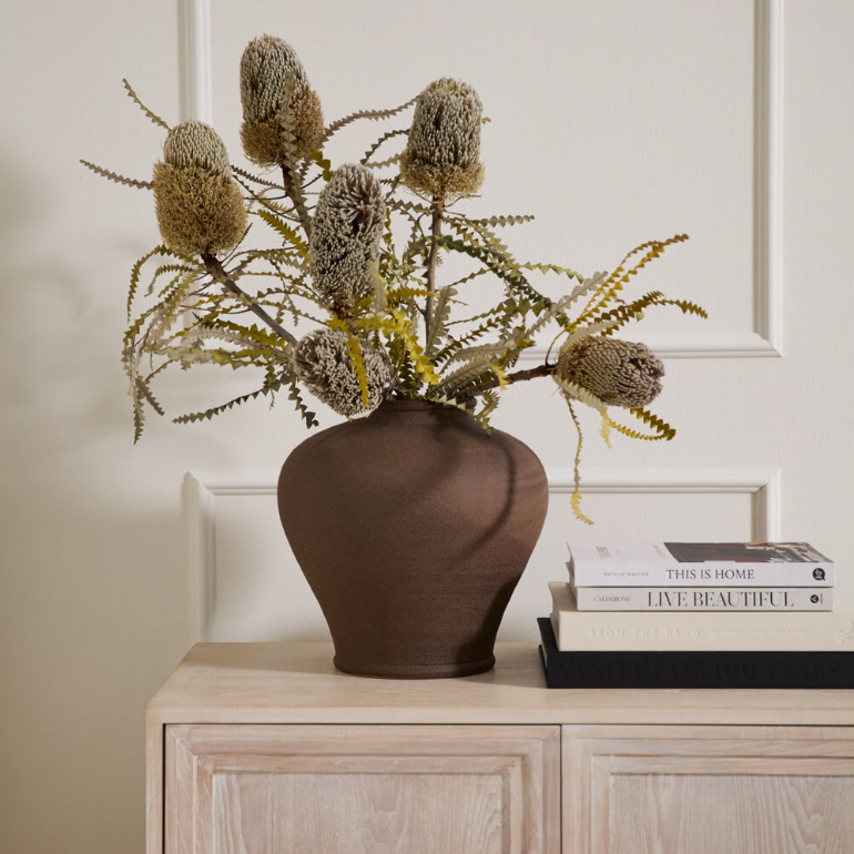 Decorative vase with dried flowers on a wooden cabinet against a neutral wall.