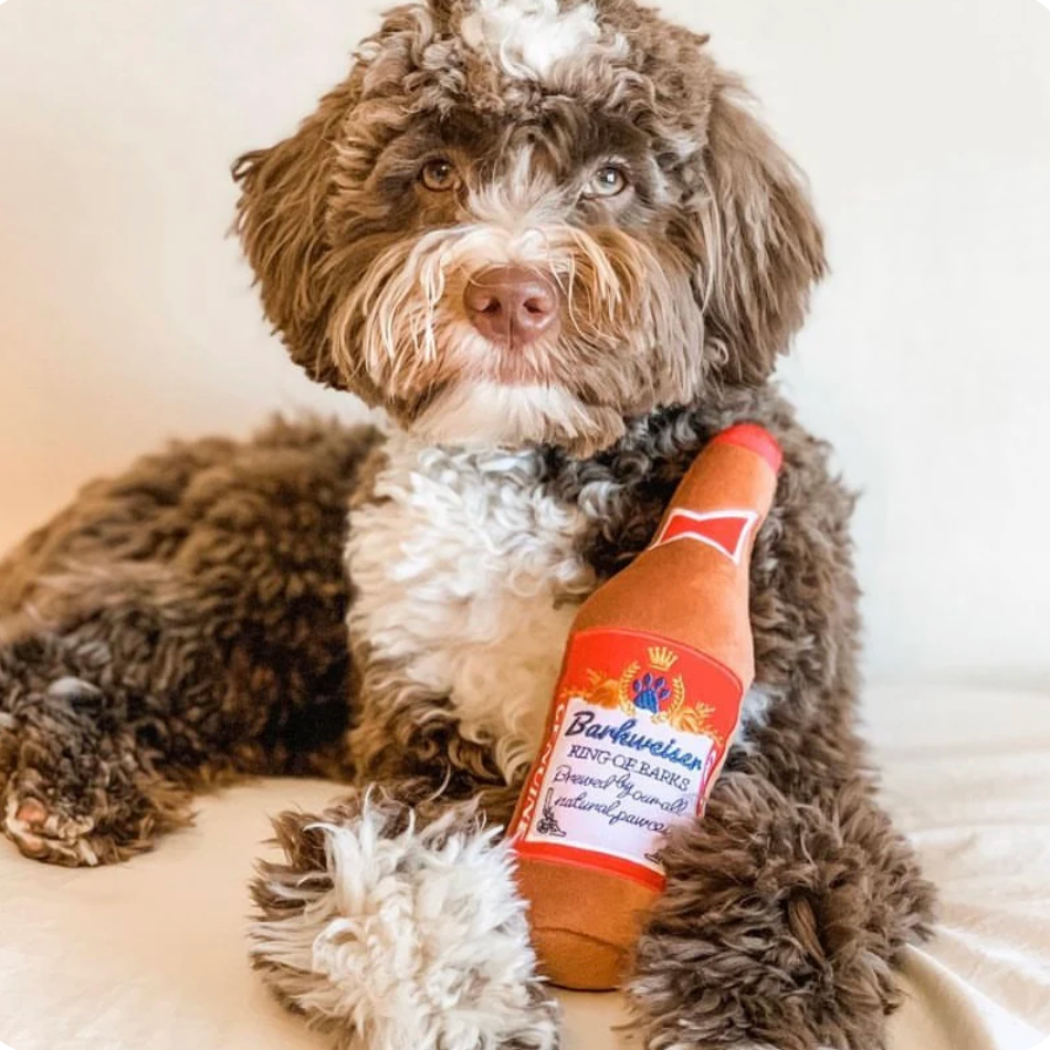 golden doodle holding Plush Barkweiser Beer Bottle dog toy with embroidered “King of Barks” label and hidden squeaker inside.
