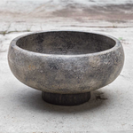 Round terracotta bowl with an aged black and gold finish, set on a woven water hyacinth base, styled as a decorative centerpiece on a concrete background