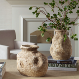 Two textured terracotta vases with an aged, rustic finish are displayed on a coffee table in a neutral-toned living room. One vase is filled with greenery, while the other sits empty beside it. The vases rest atop stacked design books, adding an organic, earthy accent to the cozy, modern space