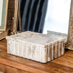 Closed rectangular marble box with scalloped lid and ribbed sides, crafted from warm-toned leopard marble, displayed on a wooden surface in front of decorative mirrors.