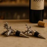 one deer shaped and one moose shaped wine bottle stoppers on a wooden surface with a blurred wine bottle in the background.