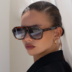 A close-up of a model wearing oversized square sunglasses with a tortoiseshell frame and dark gradient lenses, styled with gold earrings and a black top against a soft white background.