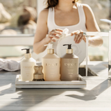 Three L'AVANT Lifestyle soap and shampoo bottles in cream, beige, and taupe tones with gold caps, arranged on a white tray on a sunlit kitchen counter, with a woman in the background washing a white plate.