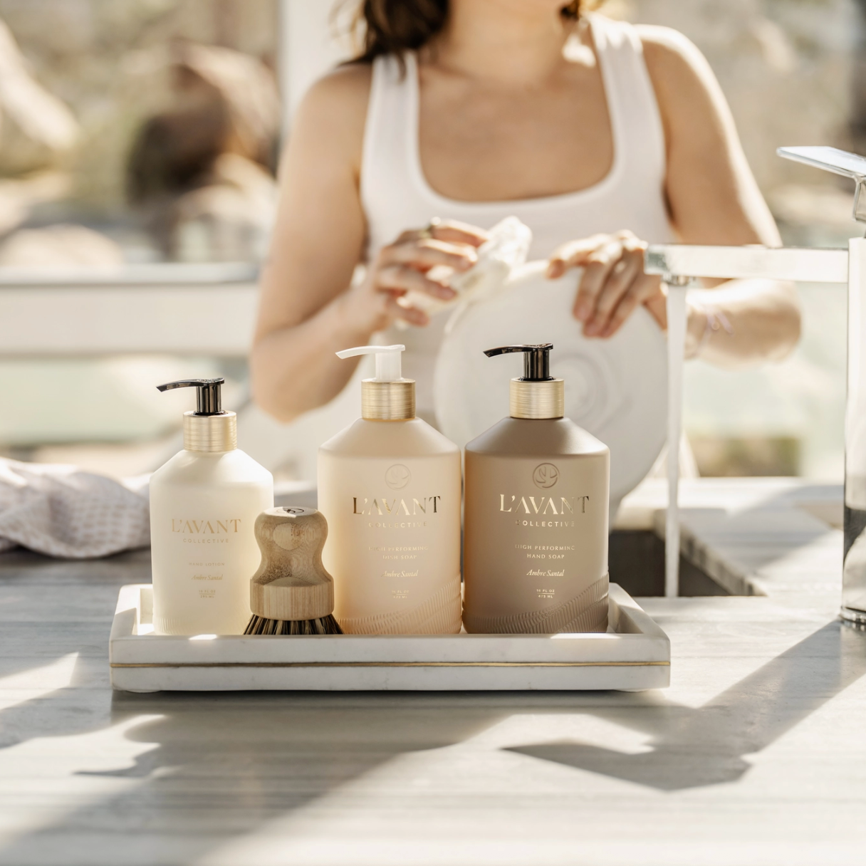 Three Lavant products in frosted beige bottles with gold pumps are arranged on a white tray on a bathroom counter, with a woman washing her hands in the softly blurred background.