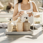 Three Lavant products in frosted beige bottles with gold pumps are arranged on a white tray on a bathroom counter, with a woman washing her hands in the softly blurred background.