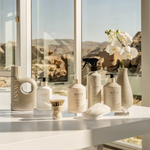A collection of LAVANT luxury bath and body products arranged on a white marble bathroom counter with a window view of a rocky desert landscape. The products include various bottles and a spray in neutral beige, white, and black tones, styled with a small vase of white orchids, a scrubber, and a sponge.
