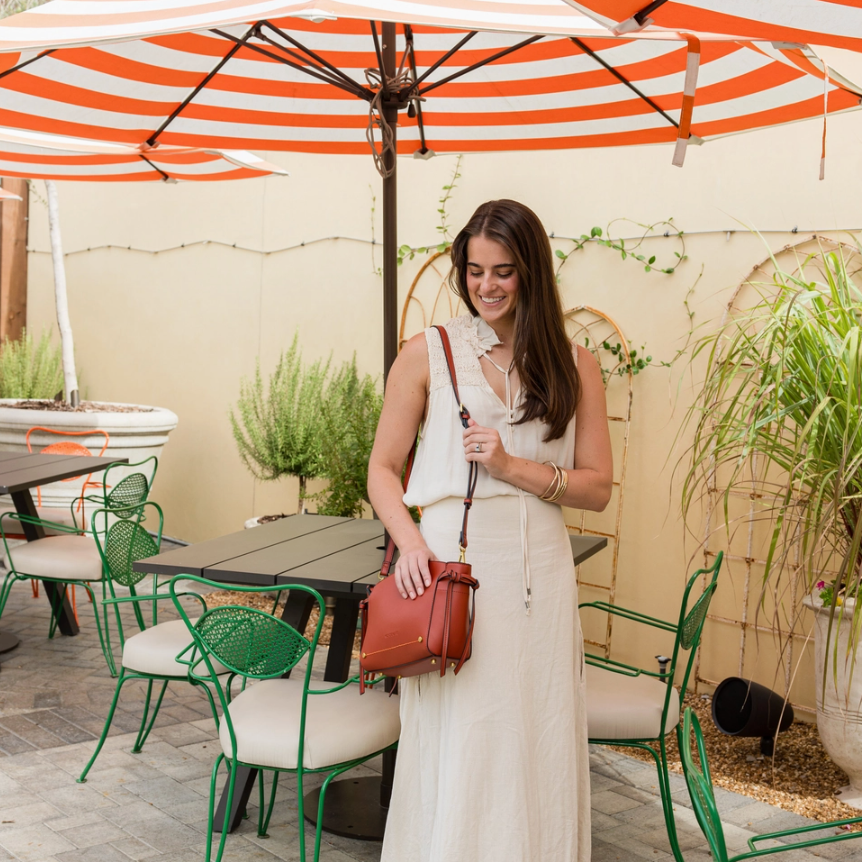 A woman stands on an outdoor patio under a large orange and white striped umbrella, wearing a sleeveless cream maxi dress and carrying a brown leather crossbody bag.