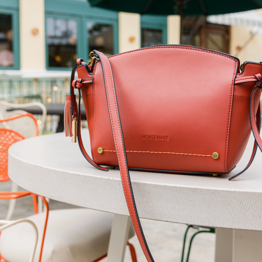 A terracotta red MONTEMARE Roma leather shoulder bag sits on a white outdoor patio table. The bag features gold-tone hardware, a matching leather strap, and a small brand logo embossed on the front.