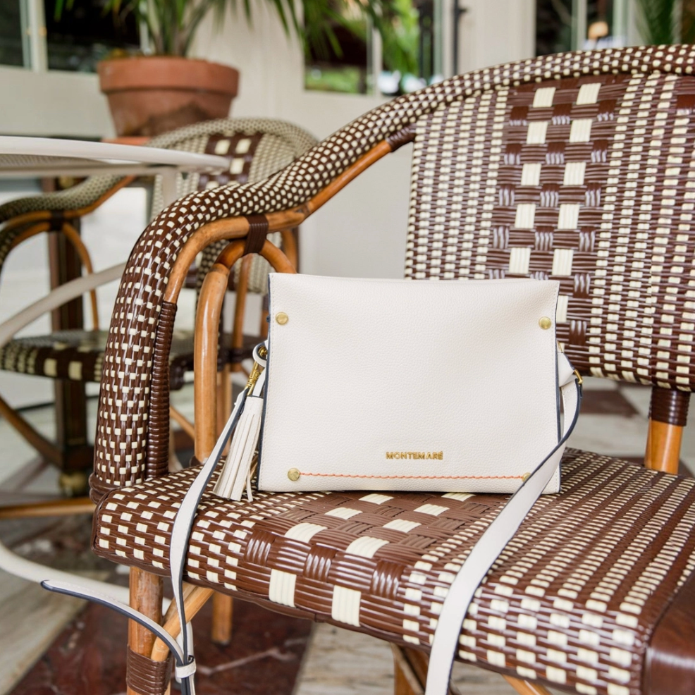 A white leather Montemare Vienna Crossbody handbag with gold hardware and tassel straps rests on a brown and cream woven rattan patio chair in an outdoor patio setting.