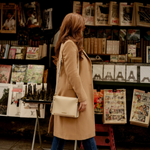 A woman in a camel trench coat and blue jeans stands in front of a street newspaper stand, carrying a cream-colored Montemare Vienna Crossbody bag with a gold logo.