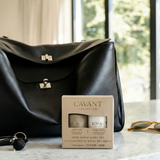A black leather handbag sits on a marble countertop in a bright room, next to a boxed Lavant Collection Mini Hand Care Set. A pair of sunglasses and a key are also visible on the counter.