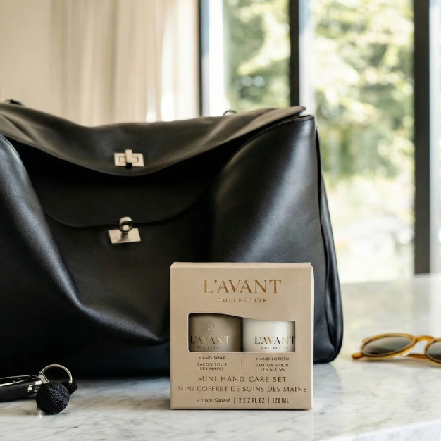 A black leather handbag sits on a marble countertop in a bright room, next to a boxed Lavant Collection Mini Hand Care Set. A pair of sunglasses and a key are also visible on the counter.