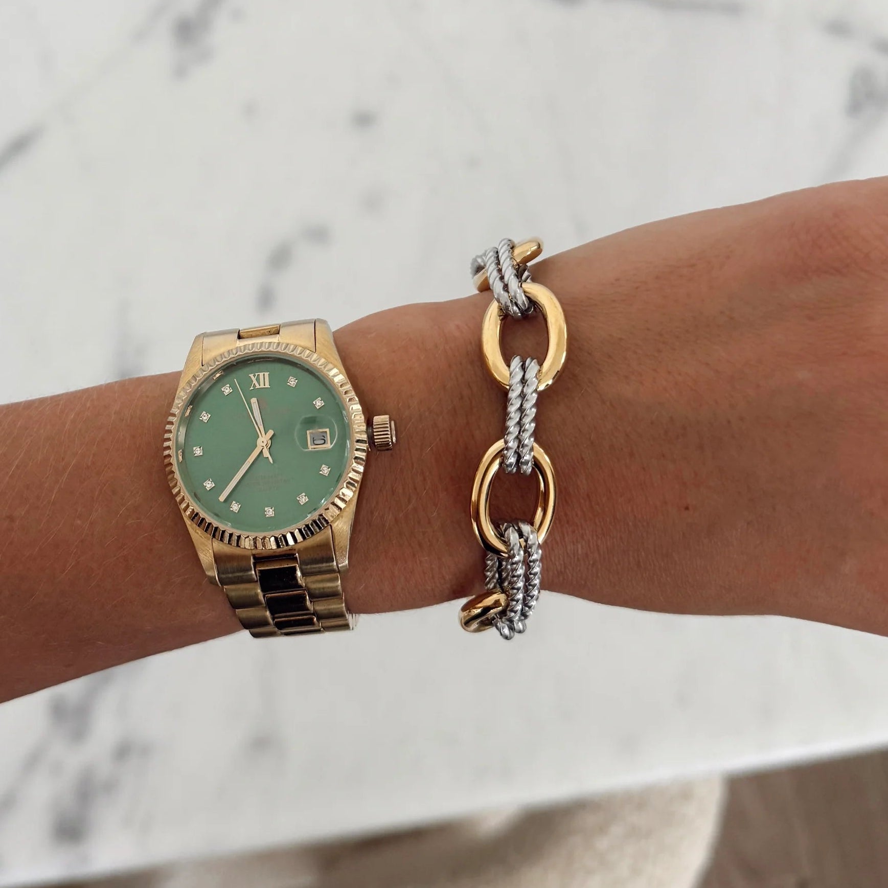 Gold watch with green face and bracelet on wrist against a marble background