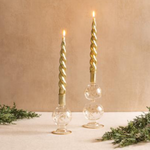 Two gold twisted candles in glass holders on a beige background with greenery.